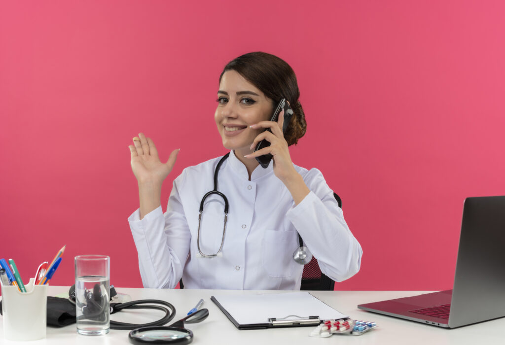 smiling young female doctor wearing medical robe with stethoscope sitting at desk work on computer with medical tools speaks on phone